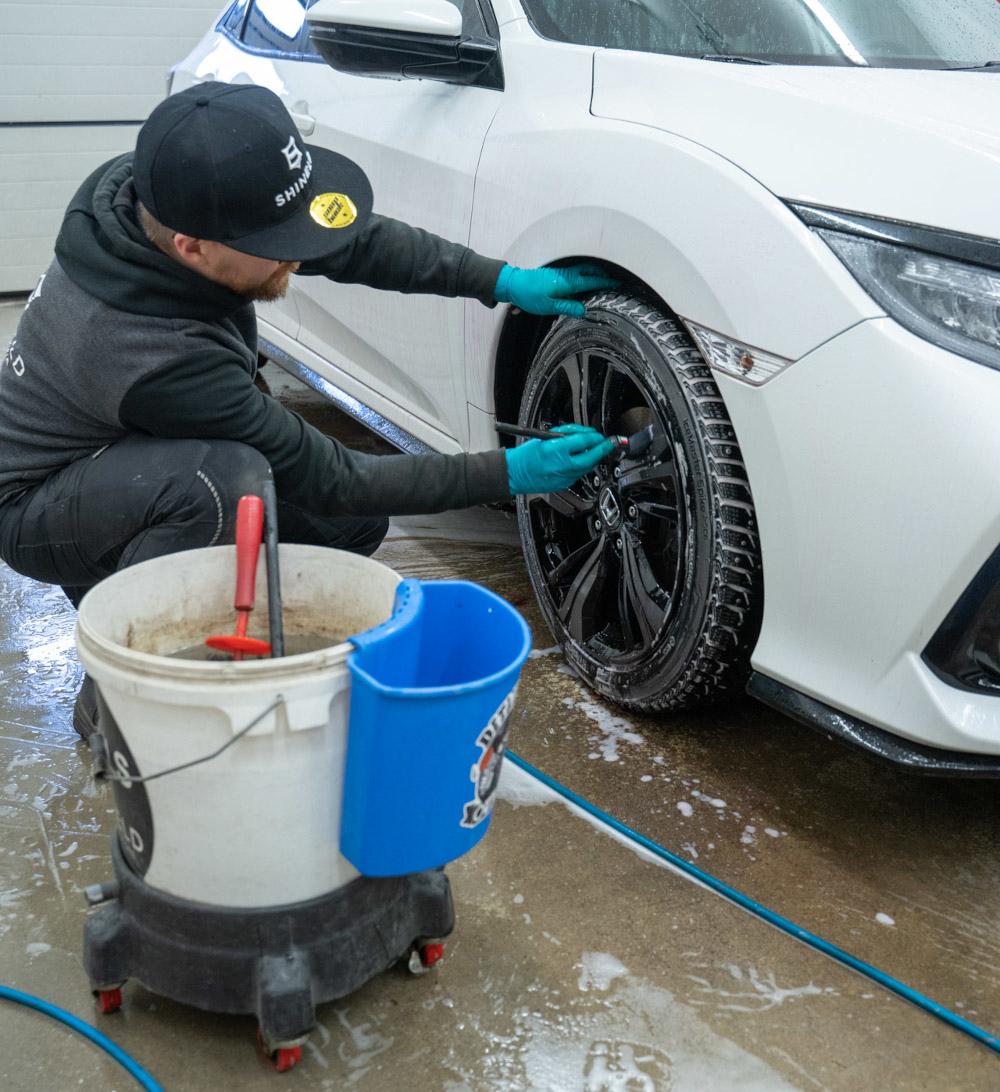 Kuvassa mieshenkilö pesemässä auton vannetta harjalla. Kuvassa näkyy autofiksari tummissa vaatteissa, valkoinen auto jossa tummat vanteet, pesuämpäri sekä vannepesu-ainetta ja -välineitä. Lattialla näkyy valkoista vaahtoa.